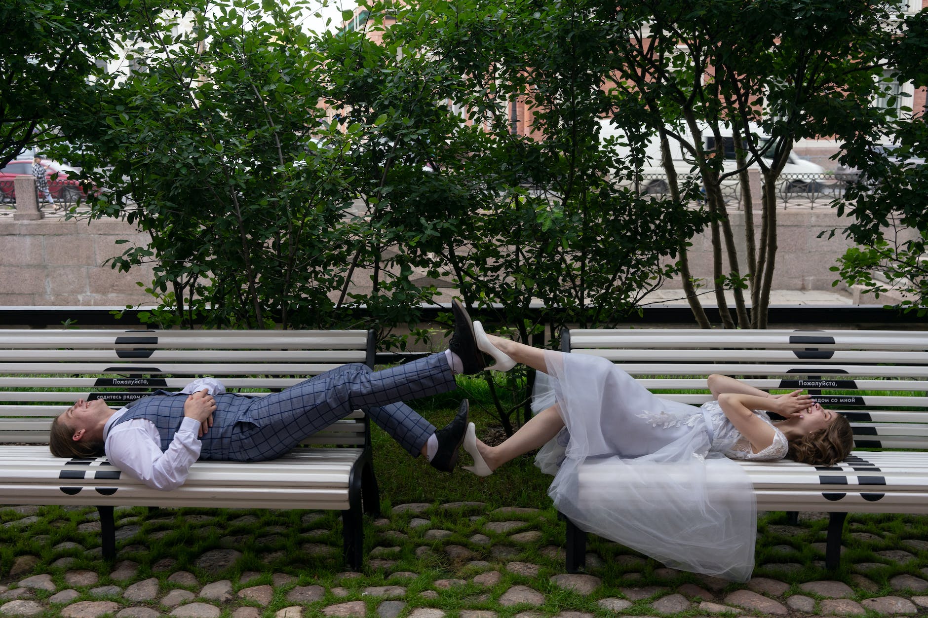 smiling newlywed couple touching legs while lying on street benches